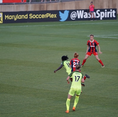 A soccer match taking place on a grassy field, featuring four players actively involved in the game. The players are wearing brightly colored jerseys, with one team in neon green and the other in red and blue. Advertising boards are visible in the background, indicating the location as Maryland SoccerPlex.