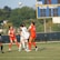 A group of female soccer players are actively engaged in a game on an outdoor field. The players are wearing red and white uniforms, with one goalkeeper in black. A soccer ball is airborne above them, indicating action and movement. A scoreboard in the background shows the score and other game details.